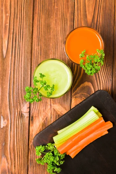 Carrot and celery juice in glasses with cut fresh vegetables and green parsley leaves on kitchen board on wooden background - top view of fresh raw healthy organic vegetarian food.