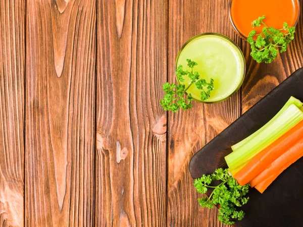 Carrot and celery juice in glasses with cut fresh vegetables and green parsley leaves on kitchen board with copy space on wooden background - top view of fresh raw healthy organic vegetarian food.