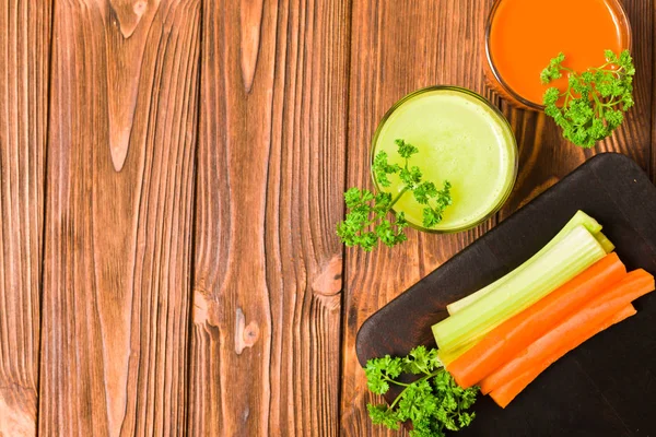 Carrot and celery juice in glasses with cut fresh vegetables and green parsley leaves on kitchen board with copy space on wooden background - top view of fresh raw healthy organic vegetarian food.
