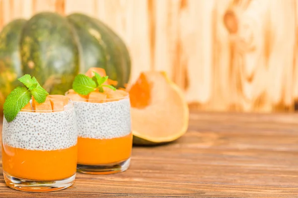 Chia pudding with pumpkin puree in beautiful glasses with green mint leaves and fresh ripe vegetable on brown wooden background - raw vegetarian sweet healthy organic dessert.