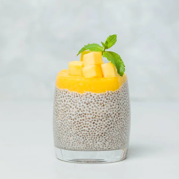 Chia seeds pudding with mango puree in beautiful glasses with green mint leaves and cut fresh ripe tropical fruit on light gray background - raw vegetarian sweet organic dessert.