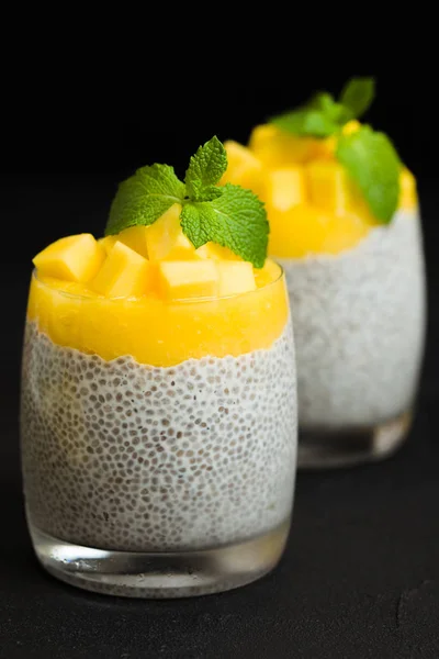 Chia seeds pudding with mango puree in beautiful glasses with green mint leaves and cut fresh ripe tropical fruit on black background - raw vegetarian sweet organic dessert in dark mood style.
