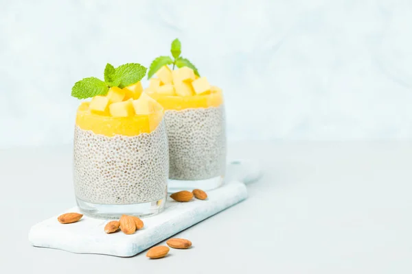 Chia seeds pudding from almond milk with mango puree in beautiful glasses with green mint leaves and cut fresh ripe tropical fruit on pastel blue background - raw vegetarian sweet organic dessert.