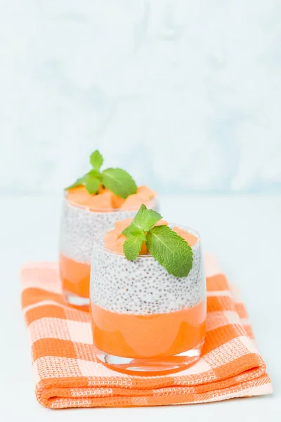 Chia seeds pudding with pumpkin puree in beautiful glasses with green mint leaves and cut fresh ripe orange vegetable on pastel blue background - raw vegetarian sweet organic dessert.