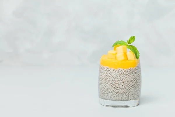 Chia seeds pudding with mango puree in beautiful glasses with green mint leaves and cut fresh ripe tropical fruit on light gray background - raw vegetarian sweet organic dessert.