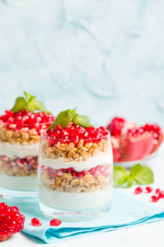 Pomegranate parfait - sweet organic layered dessert with granola flakes, yogurt and ripe fruit seeds in beautiful glasses on pastel blue background with copy space. Natural vegetarian healthy food.