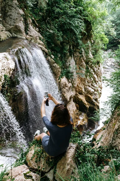 Bir gezgin bir telefonla Agur gorge büyük bir şelale fotoğrafını bir uçurumun kenarında alır.