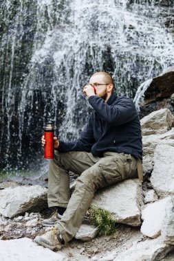 Genç bir adam üzerinden bir şelale yakınındaki taş üzerinde oturan bir termos çay veya kahve içer