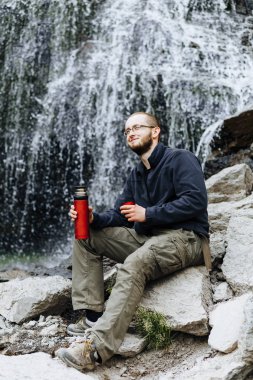 Genç bir adam üzerinden bir şelale yakınındaki taş üzerinde oturan bir termos çay veya kahve içer
