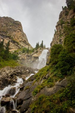 Yüksek Şelale, akan nehir, arka planda yüksek dağ olan akarsu. Kayalar, uçurumlar, yeşil çimenler, orman ağaçları. Kuzey manzara fotoğrafı.