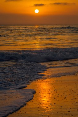 Biraz bulutlu bir günde günbatımı üzerinde bir beach Carlsbad, California. Dalgalar kıyı yıkama turuncu gökyüzü ve yansıma şunlardır. Küçük, köpüklü dalgalar ön planda kırma vardır. Uzak bir gemi ufukta yüzen.