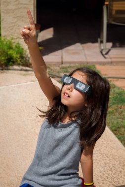 A young girl, with brown hair, sits on a ball outside on a bright, sunny day.  She is wearing eclipse glasses, paper glasses with solar filters for lenses, and pointing to the sky with a look of wonder. clipart