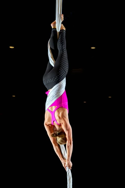 A female aerialist with a muscular back spins upside down in a skater pose against a black background.  She is wearing a bright pink athletic shirt and polkadot leggings.  Her silk is white and you she is lit in a way to show off her muscles.