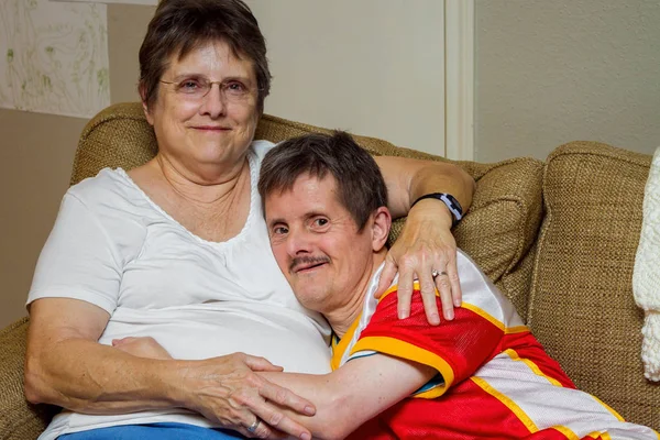 An older man with Downs Syndrome, hugs his older sister as they sit on a couch. The woman looks tired, the man looks mischeivious.  He is about to tickle her.