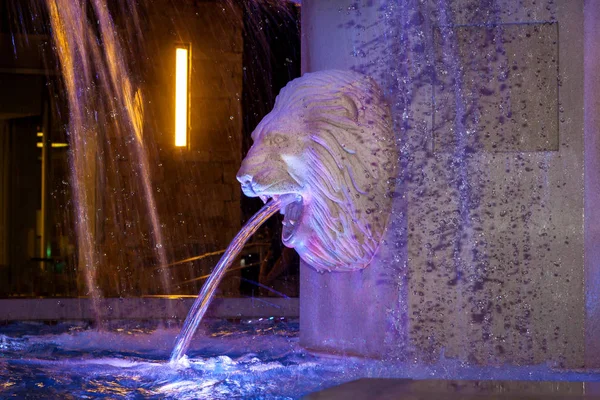 Closeup of a lion head on a water fountain.  The lion is spitting out water which is slightly blurred due to motion.  It is nighttime and the fountain is lit with purple light.  Some of the water motion is stopped by the flash.