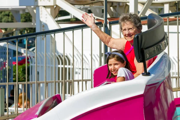 An elderly woman rides on a tilting, spinning, whirling ride at a theme park with her young granddaughter.  Both are smiling and the grandmother is waving at the camera as their car spins around.