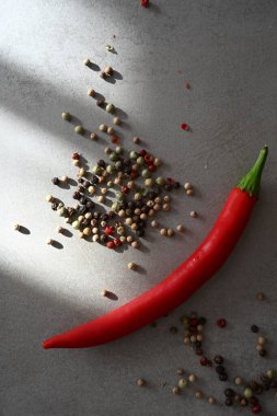 Canlı bir kırmızı biber gri bir yüzey üzerinde siyah, beyaz ve yeşil biber karışımının yanında yer alır. Işık, baharatların dokularını ve zengin renklerini vurgular..