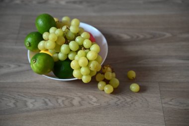 Ahşap bir yüzeyde beyaz bir kase yeşil üzüm, parlak limon ve diğer meyvelerle dolup taşıyor. Kasenin etrafına saçılmış birkaç üzüm, doğal ve sağlıklı hissi ön plana çıkarıyor..
