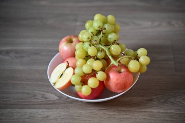 Tahta bir yüzeyde beyaz bir kase kırmızı elma ve yeşil üzümlerin taze bir karışımını barındırır. Bir elma dilimlenir ve çekirdeğini ortaya çıkarır. Meyvenin taze ve sağlıklı karakterini ön plana çıkarır..
