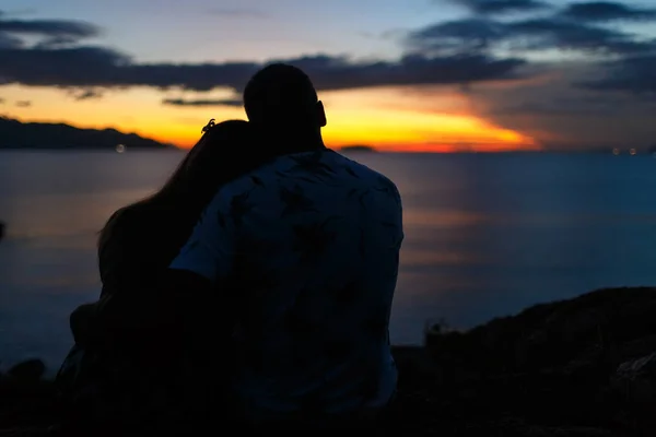 Silhouette of a couple are sitting and hugging against the background of the sea and sunrise. The girl's head on the guy's shoulder. Back view.