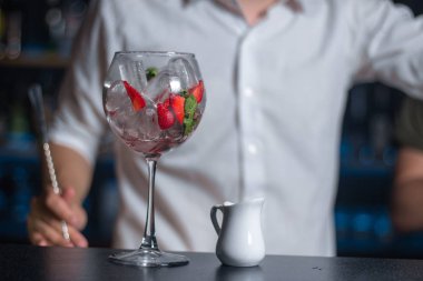 Şarap kadehi buz, çilek ve nane ile dolu, tezgahtaki beyaz süt sürahisinin yanında. Arka planda bulanık barmen. Kokteyl hazırlama süreci