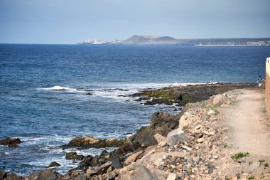 Lüks sahil. Kayalık sahil okyanusun dalgalar kayalara karşı yendi. Bir turist mavi bir sırt çantası ile arka planda elini kolunu sallaya dönüyor. Şehrin liman ile uzaktan görülür.