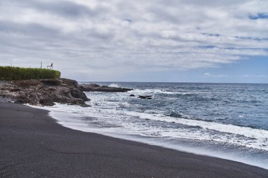 Siyah kum ile olağandışı plaj. Güzel temiz plaj sıcak deniz, okyanus. Güneşlenmek için zamanı. Boş tesisi alanı. Volkanik kayalar suya yakın. Dalgalar sıçrama sahile hakkında.