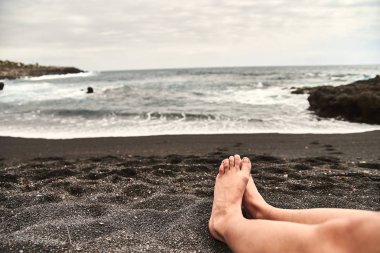 Sahilde kadın bacaklar. Siyah volkanik kum ile benzersiz bir plaj. Dalgalar kıyıya hakkında sıçrama, taze deniz meltemi esiyor. Kadın, kız sahilde bir bulutlu gün dinlenme.
