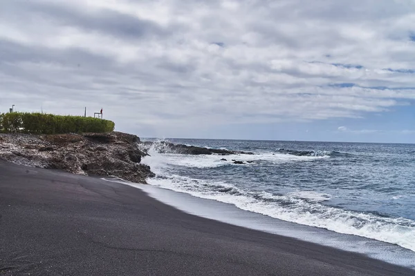 stock image Unusual beach with black sand. Beautiful clean beach with warm sea, ocean. It's time to sunbathe. Empty resort area. Volcanic rock near the water. Waves splash about the shore.