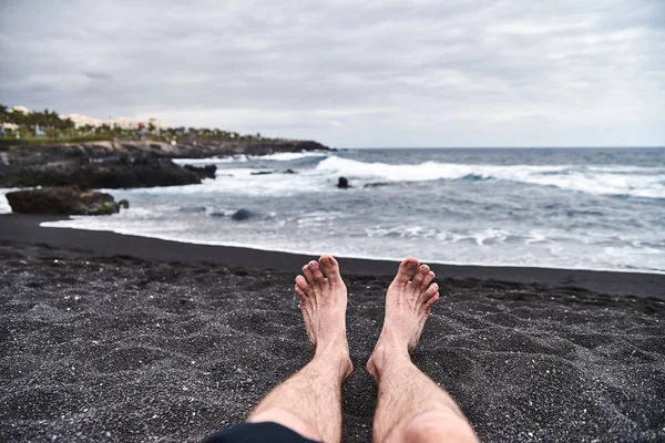 Men\'s legs on the beach. A unique beach with black volcanic sand. Waves splash about the shore, a fresh sea breeze is blowing. A man, a guy, a boy is resting on the beach.