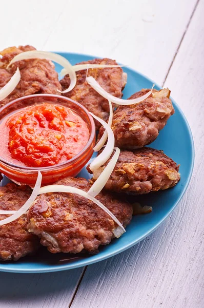 Buffet served in a hearty breakfast, lunch and dinner. Appetizing meat cutlet with fresh onions on a blue plate. Tomato-pepper sauce for meat. A plate of food on a white wooden table.