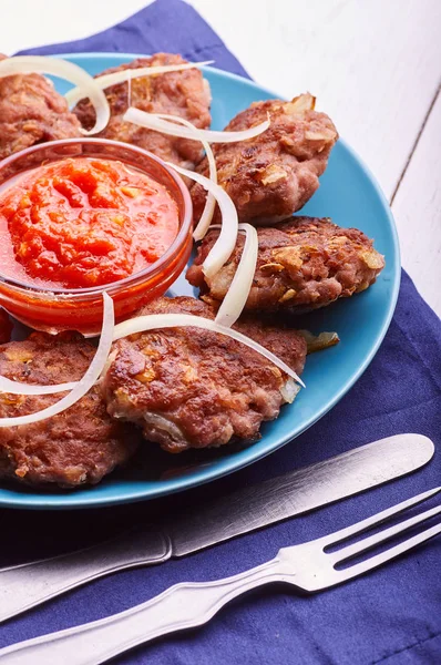Buffet served in a hearty breakfast, lunch and dinner. Appetizing meat cutlet with fresh onions on a blue plate. Tomato-pepper sauce for meat. Blue napkin and cutlery on a white wooden table.