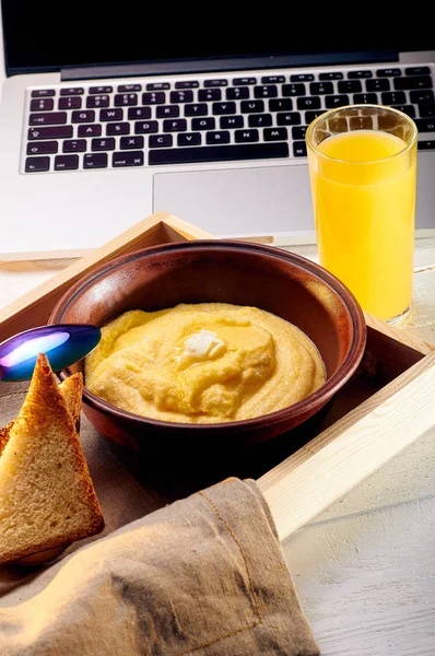 笔记本电脑附近有一份阳光明媚的早餐 玉米粥配黄油和一些脆面包 橙色新鲜榨汁和食物在托盘上的早餐在床上 — 图库照片