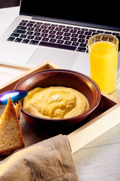 笔记本电脑附近有一份阳光明媚的早餐 玉米粥配黄油和一些脆面包 橙色新鲜榨汁和食物在托盘上的早餐在床上 — 图库照片