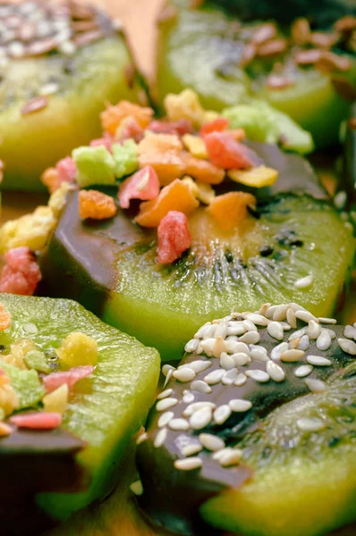 A bright fruit dessert for vegetarians and slimming. Kiwi in chocolate is decorated with small colored candied fruits, flax seeds and sesame seeds.