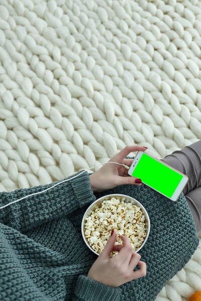 Very cozy picture for the autumn, September, October, November. A girl is watching movie on phone with headphones and eating popcorn. Green screen on your mobile for your picture, text, information.