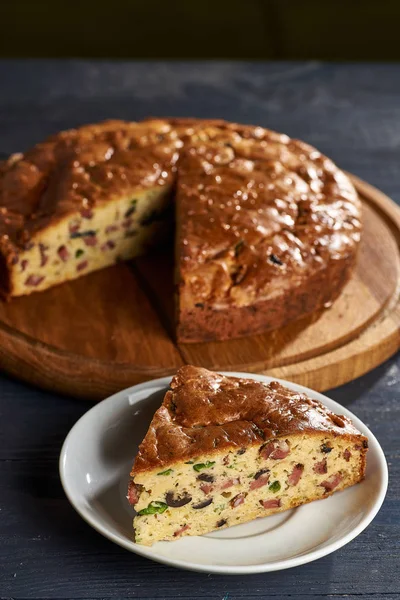 A piece of unusual unsweetened pie in the foreground. Corn wheat pie with various options of salty stuffing in the background. Snack cake with sausage, olives, vegetables, greens, meat.