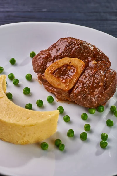 Unusual serving of food in a cafe, restaurant. Potato puree / polenta in the form of an incomplete moon, green peas in the form of stars in the sky. A large piece of appetizing meat on the bone.