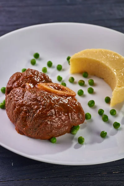 Unusual serving of food in a cafe, restaurant. Potato puree / polenta in the form of an incomplete moon, green peas in the form of stars in the sky. A large piece of appetizing meat on the bone.