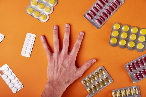 A man chooses medicine for treatment. Male hand on an orange background and a lot of different pills, drugs, vitamins, minerals, food additives. The doctor is trying to make a diagnosis.