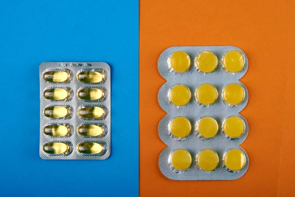 Two types of yellow pills on two-tone blue-orange background. Fish oil, omega-3 and sucking candies from sore throat. Oblong translucent gelatin-coated capsules and classic round sucking tablets.
