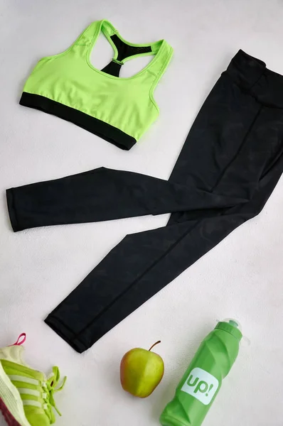 Sport time. Sports flat lay in green tones. Black leggings and green top on a white background. Everything you need for a healthy lifestyle. Clothes, sneakers, apple and a fashionable flask for water.