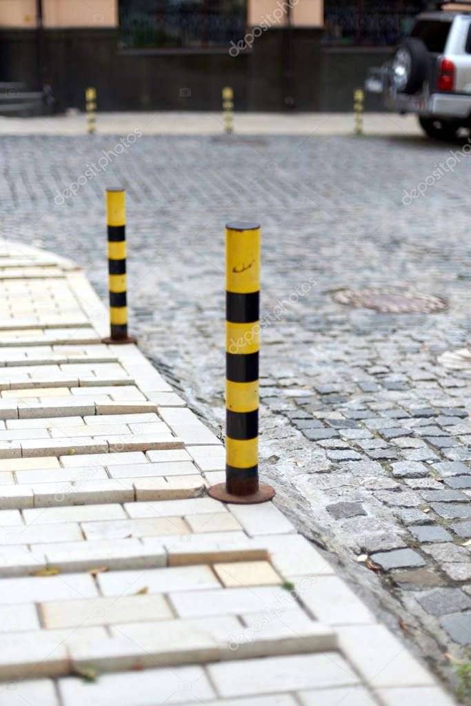 Restrictive posts between the pedestrian sidewalk and the road. Small low poles of yellow-black color, striped. Beautiful old road in Europe, paved with stone blocks. Cozy street.
