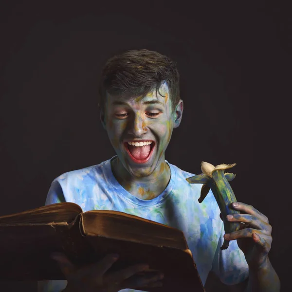 young interesting guy painted with colorful paints with a happy expression reads a book and eats a painted banana