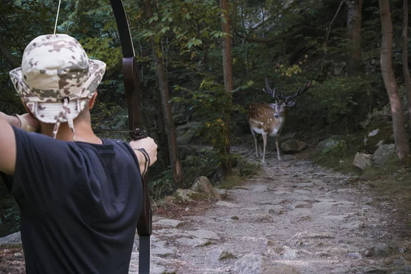 hunter with an arrow aimed at a spotted deer on a narrow mountain path