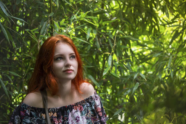 Young beautiful red-haired girl among the green bamboo forest looks at the sunlight through the foliage