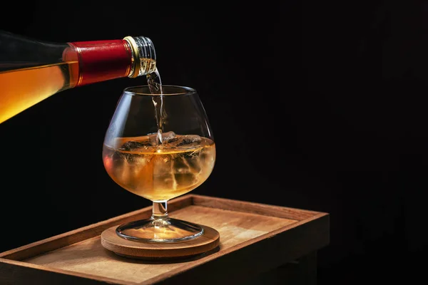 Whiskey is poured into a brandy glass with ice, the glass is ful — Stock Photo, Image