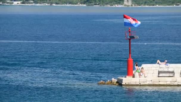 Molo di pietra con faro e bandiera battente alla luce del giorno, persone che camminano e si siedono su un muro di pietra — Video Stock