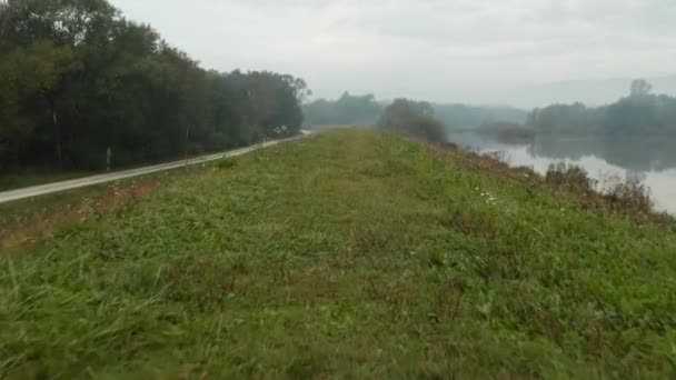 Drohnenschuss, der an einem nebligen Herbstmorgen entlang eines grasbewachsenen Hügels mit einem nahe gelegenen spiegelnden See fliegt, — Stockvideo