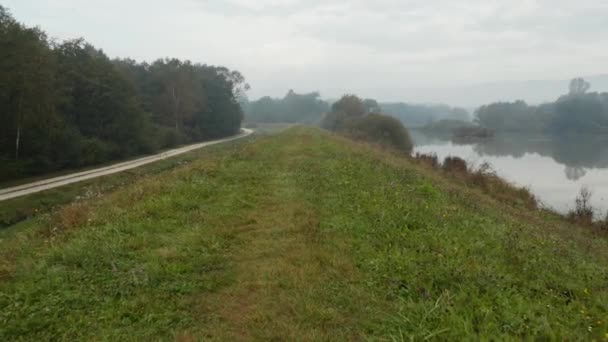 Drohnenschuss, der an einem nebligen Herbstmorgen entlang eines grasbewachsenen Hügels mit einem nahe gelegenen spiegelnden See fliegt — Stockvideo